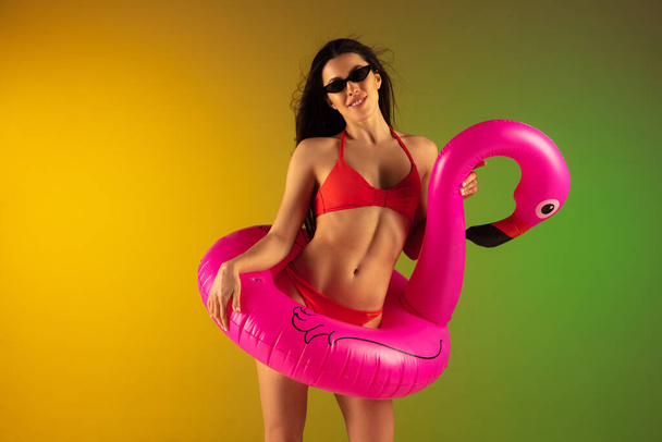 Fashion portrait of young fit and sportive woman in stylish red luxury swimwear with rubber flamingo on gradient background. Perfect body ready for summertime. - Foto, Imagem