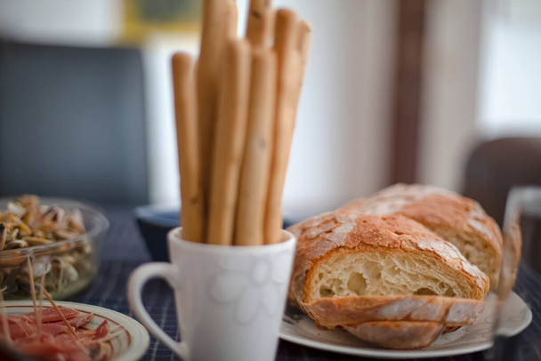 Vista de la mesa preparada para el almuerzo, con pan rústico en rodajas, taza con palitos de pan tostados y rebanadas de jamón
... - Foto, imagen
