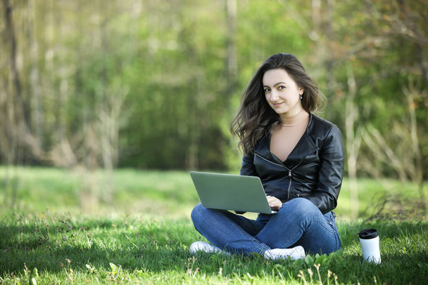 Young woman in a black jacked with coffee cup and laptop working outside in a park. Remote work. Digital work. Distance learning - Photo, Image