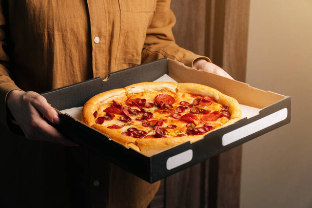 Women's hands hold a beautiful delicious pepperoni pizza in front of the door. Food delivery to the office. - Foto, Imagen