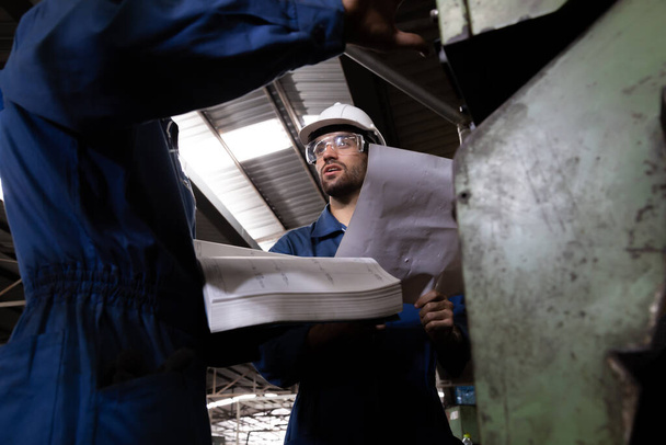 Инженер-заводчик читает инструкцию по эксплуатации машины и стоит с голубым рабочим комплектом одежды и защитным шлемом. Технические специалисты-механики на заводе тяжелой промышленности
 - Фото, изображение