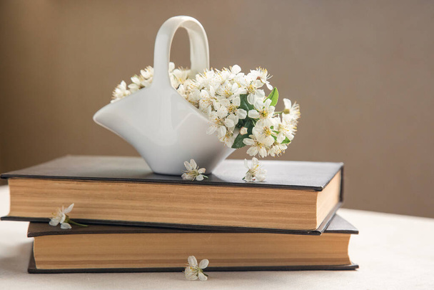 Mały wazon utworzony biały kosz ceramiczny z kwiatami wiśni jest w książkach. Kwiat Wiosny. Romantyczna martwa natura w beżowych kolorach. Widok z przodu. - Zdjęcie, obraz
