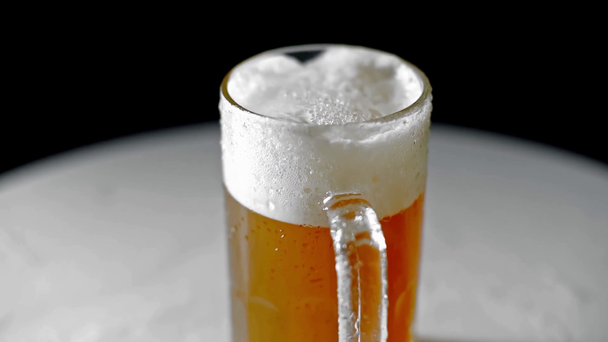 taza llena de cerveza fría con espuma, girando sobre la superficie blanca aislado en negro
 - Imágenes, Vídeo