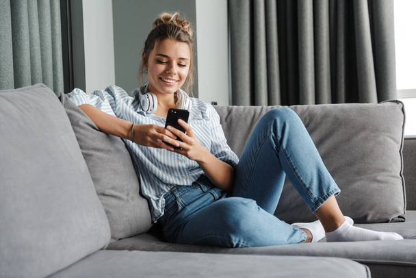Image of smiling nice woman with headphones using cellphone while sitting on sofa at living room - Фото, изображение
