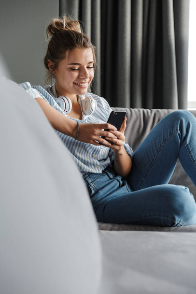 Image of smiling nice woman with headphones using cellphone while sitting on sofa at living room - Photo, Image