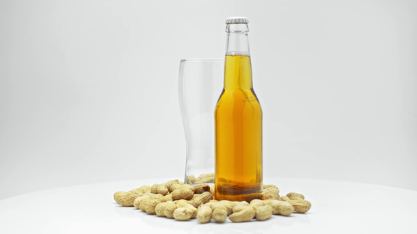 prázdný hrnek a láhev lehkého piva poblíž arašídů izolovaných na bílém - Záběry, video