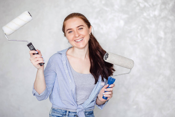 A brunette girl is resting while repairing a house. A young smiling beautiful woman in casual clothes holds a paint roller for a wall painting isolated on a gray background.  - Valokuva, kuva