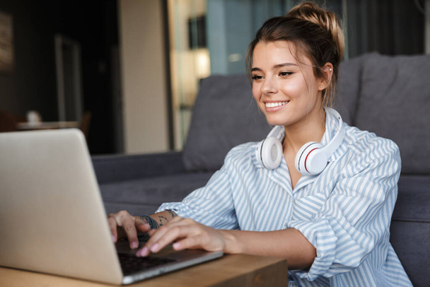 Image of smiling nice woman with headphones using laptop while sitting on floor at living room - Foto, immagini