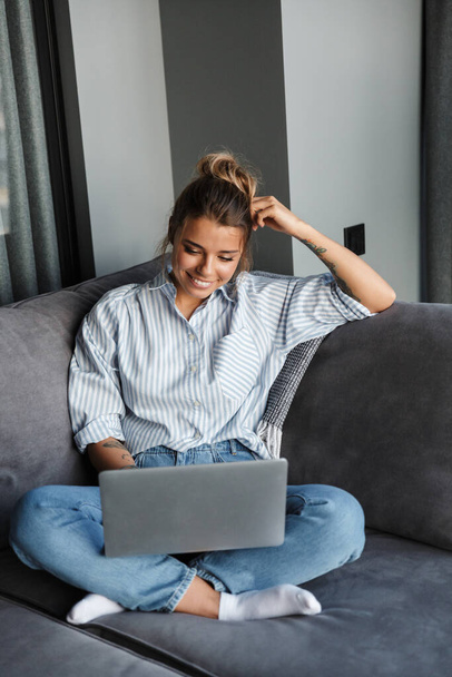 Image of joyful nice woman smiling and using laptop while sitting on couch at living room - Fotoğraf, Görsel
