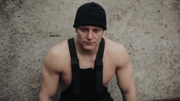 Ein junger traurig enttäuschter muskulöser Mann sitzt mit schwarzem Hut und schwarzen Overalls auf seinem nackten Körper vor einer grauen Wand. Nahaufnahme - Filmmaterial, Video