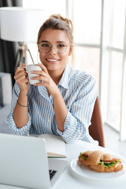 Image of joyful nice woman in eyeglasses drinking coffee and using laptop while sitting at table in living room - Фото, зображення