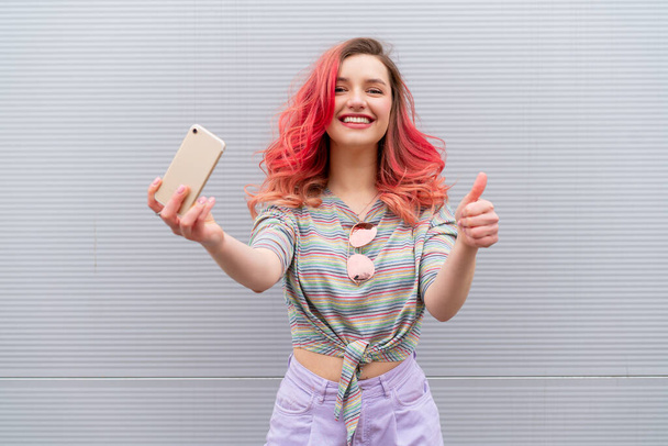 Fashion portrait of pretty smiling hipster woman in sunglasses and colored red hairs with smartphone against the colorful grey wall - Fotoğraf, Görsel