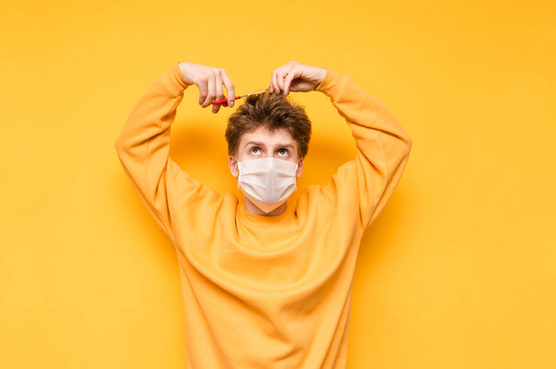 Funny guy in a medical mask cuts his hair with scissors on a yellow background. Hairdressers in quarantine. Shaggy young man cuts his own hair. Quarantined barbershop. - Photo, Image
