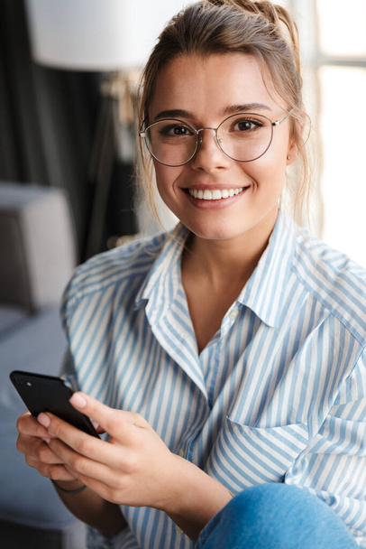 Image of happy young woman in eyeglasses smiling and using cellphone at living room - Foto, Imagem