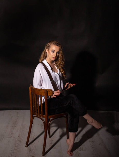 beautiful barefoot blonde in a white shirt and black pants on suspenders sits in the studio on a chair cross-legged and looking at the camera - 写真・画像