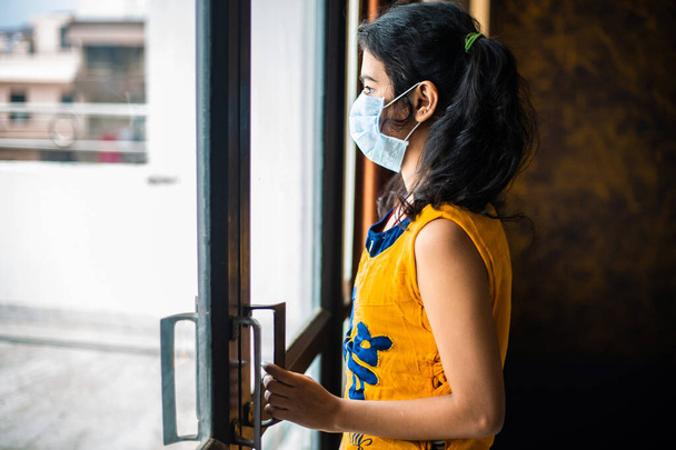 Ιός του κερατοειδούς. Άρρωστο νεαρό κορίτσι του ιού της Κορόνας κοιτάζει από το παράθυρο και φοράει μάσκα προστασίας και ανάρρωσης από την ασθένεια στο σπίτι. Καραντίνα. Ασθενής απομονωμένος στο σπίτι για την πρόληψη λοίμωξης. - Φωτογραφία, εικόνα