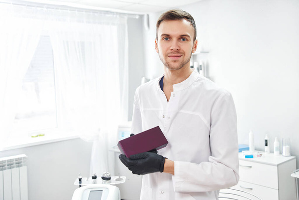 Επαγγελματίας γιατρός αισθητικός άνθρωπος σε λευκό ιατρικό παλτό χαμόγελα και κρατά το προϊόν στα χέρια, δείχνει και διαφημίζει κουτί με αντίγραφο χώρο στο παρασκήνιο του σαλόνι ομορφιάς - Φωτογραφία, εικόνα