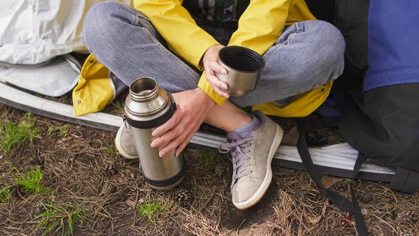 La joven turista se sienta cerca de una tienda y bebe té de thermos. Camping en el bosque - Foto, imagen