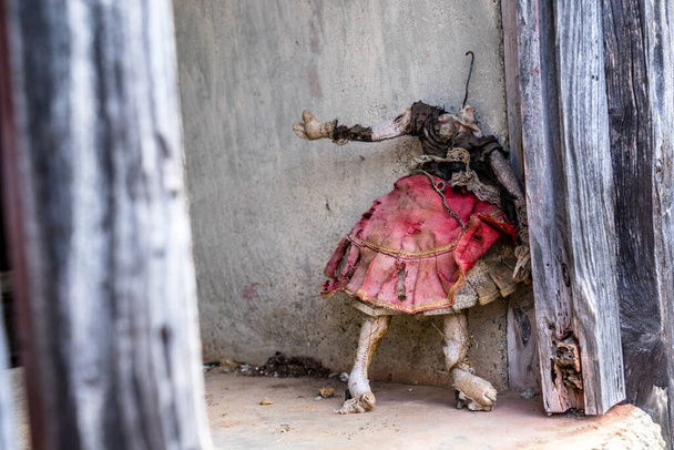muñeca vieja, sucia y sin rostro de pie en un marco de ventana en una casa abandonada en las montañas de La Gomera, Islas Canarias. Maldito, roto y malvado juguete de bebé niña en un vestido rojo desgastado y dañado
 - Foto, Imagen