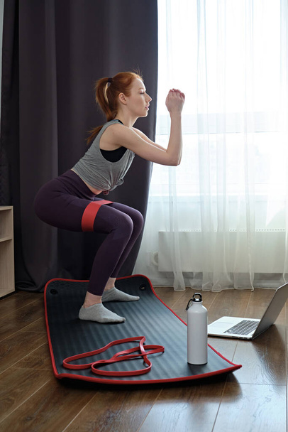 The girl squats with an elastic band on her feet, practicing at home in front of a laptop, focus n mat.  - Valokuva, kuva