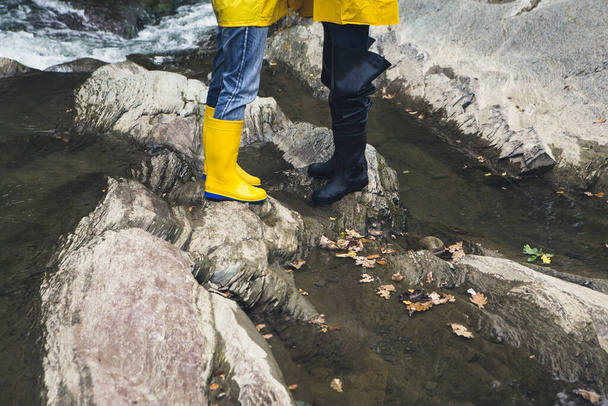 Feet of young man and woman couple in yellow raincoats and gumboots walking on rocky river  - Zdjęcie, obraz
