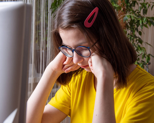 Pensive Caucasian student in a yellow T-shirt thinks, suffers, presses her hands to her face. Distance learning. Study at night. - Φωτογραφία, εικόνα