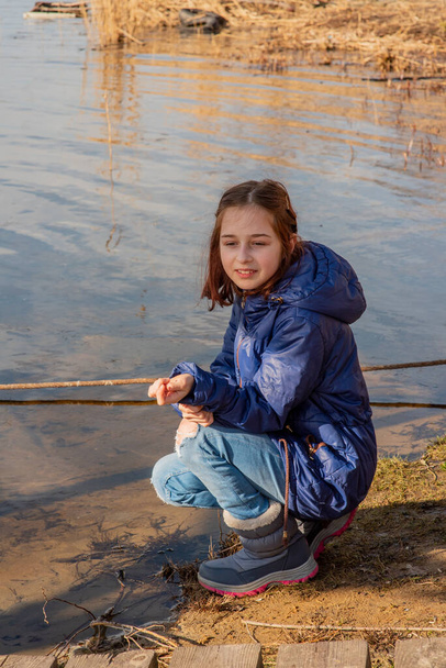 Egy könnyű báránybőrkabátos lány áll a folyó partján egy napsütéses napon. 9 éves lány, báránybőrkabátban a folyónál. 9 éves vagy 10 éves lány. Természet, gyermek, iskoláslány.tinédzser - Fotó, kép