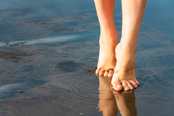 Nahaufnahme einer schlanken Frau, die am Meer spaziert. Nasser schwarzer Sand mit Reflexion der Mädchenfüße. - Foto, Bild