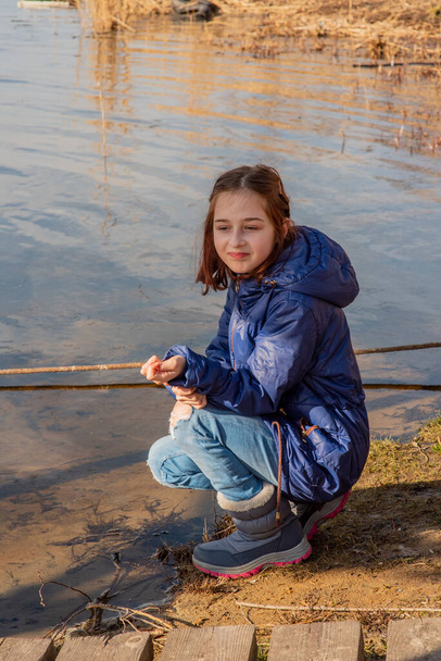 Een meisje in een lichte schapenvacht staat op een zonnige dag aan de oever van de rivier. Meisje van 9 jaar in een schapenvacht bij de rivier. Meisje van 9 of 10 jaar oud. Natuur, kind, schoolmeisje.Tiener - Foto, afbeelding