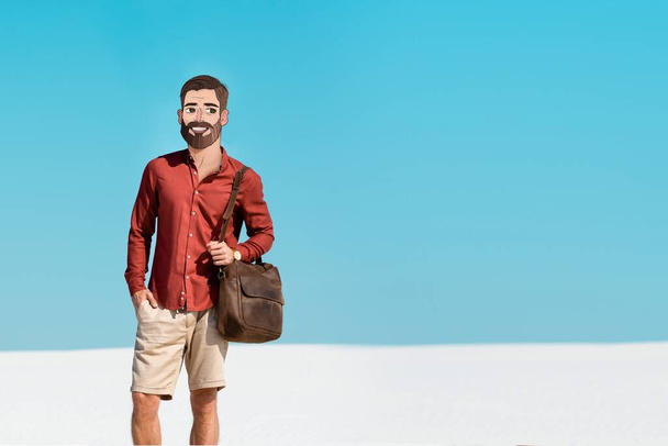 férfi mosolygós illusztrált arc gazdaság bőr táska homokos tengerparton ellen tiszta kék ég - Fotó, kép