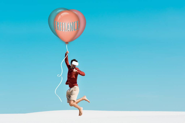 man on sandy beach in vr headset flying on balloon with freelance lettering against clear blue sky - Photo, Image