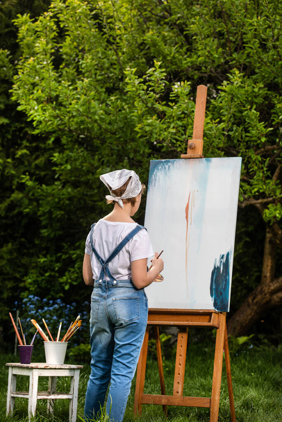 Uma jovem artista segura um pincel e pinta um quadro em um cavalete nos raios do pôr do sol. O pintor pinta pinturas a óleo ao pôr-do-sol
. - Foto, Imagem