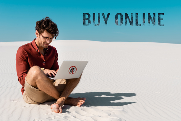 young man on sandy beach using laptop against clear blue sky, buy online illustration - Photo, Image