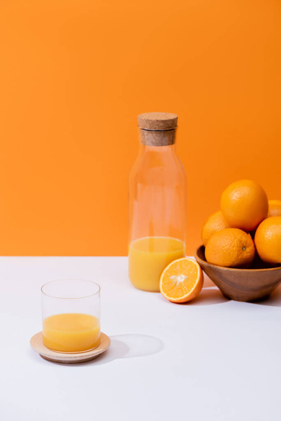 fresh orange juice in glass and bottle near oranges in bowl on white surface isolated on orange - Fotografie, Obrázek