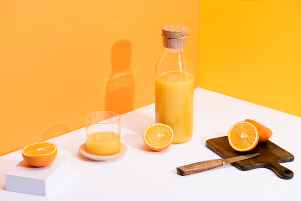 friss narancslé üvegben és palackban érett narancs közelében, fa vágódeszka késsel fehér felületen narancssárga alapon - Fotó, kép