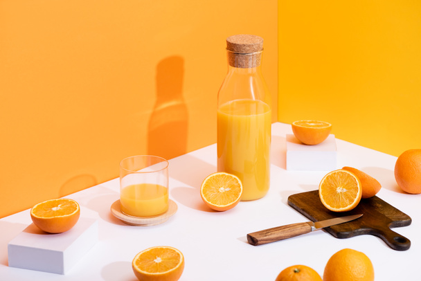 fresh orange juice in glass and bottle near ripe oranges, wooden cutting board with knife on white surface on orange background - Photo, Image