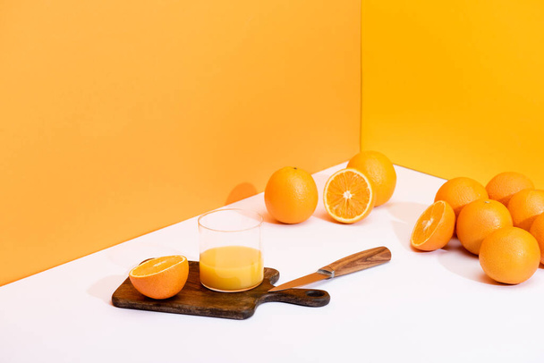 čerstvý pomerančový džus ve skle na dřevěné řezací desce s nožem v blízkosti zralých pomerančů na bílém povrchu na oranžovém pozadí - Fotografie, Obrázek