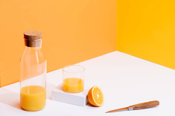 fresh orange juice in glass and bottle near ripe orange and knife on white surface on orange background - Photo, Image