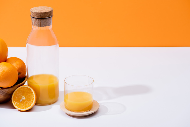 čerstvý pomerančový džus ve skle a láhvi v blízkosti pomerančů v dřevěné míse na bílém povrchu izolované na oranžové - Fotografie, Obrázek