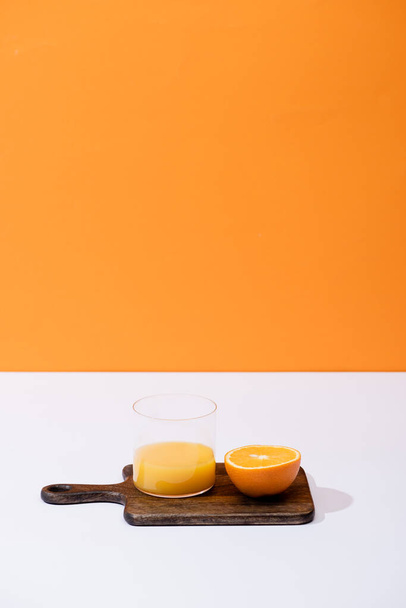 čerstvý pomerančový džus ve skle u řezaného ovoce na dřevěné řezací desce na bílém povrchu izolované na oranžové - Fotografie, Obrázek