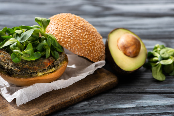 tasty vegan burger with microgreens served on cutting board near avocado on wooden table - Foto, Imagem