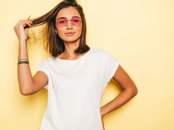 Young beautiful woman looking at camera.Trendy girl in casual summer white T-shirt and jeans shorts in round sunglasses. Positive female shows facial emotions. Funny model isolated on yellow  - Fotó, kép