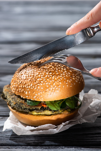 cropped view of woman cutting tasty vegan burger with fork and knife - Photo, Image