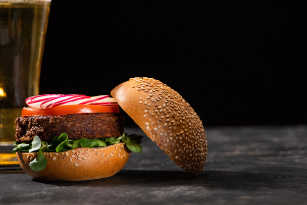 enfoque selectivo de sabrosa hamburguesa vegana con microgreens, rábano y tomate servido en superficie texturizada con cerveza aislada en negro
 - Foto, Imagen