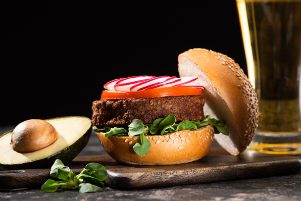selective focus of tasty vegan burger with vegetables served on wooden cutting board with avocado and beer isolated on black - 写真・画像