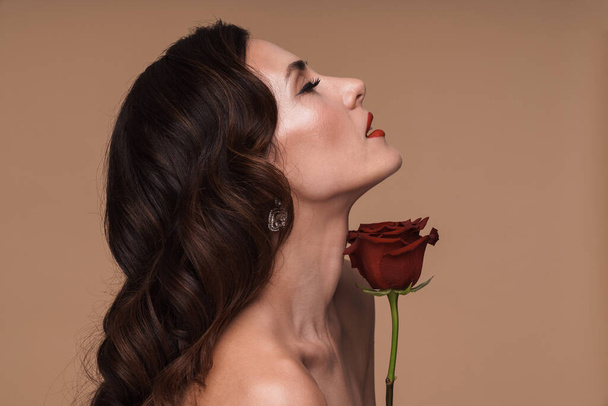 Beauty portrait of brunette gorgeous adult half-naked woman holding red rose isolated over beige background - 写真・画像
