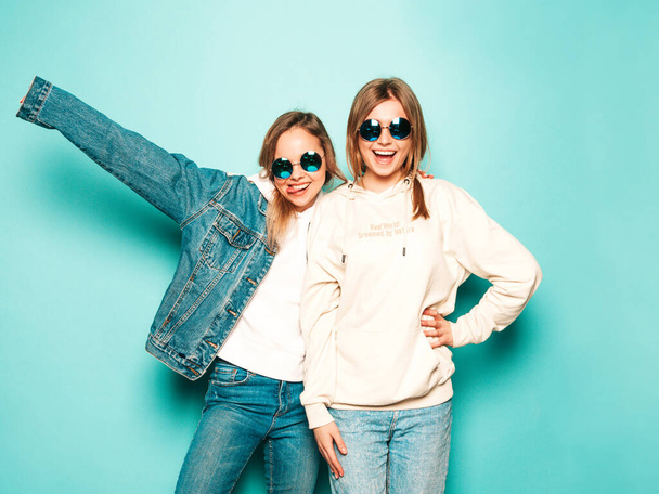 Two young beautiful brunette smiling hipster girls in trendy summer hoodie and jeans jacket clothes. Sexy carefree women posing near blue wall. Trendy and positive models having fun in sunglasses - Zdjęcie, obraz
