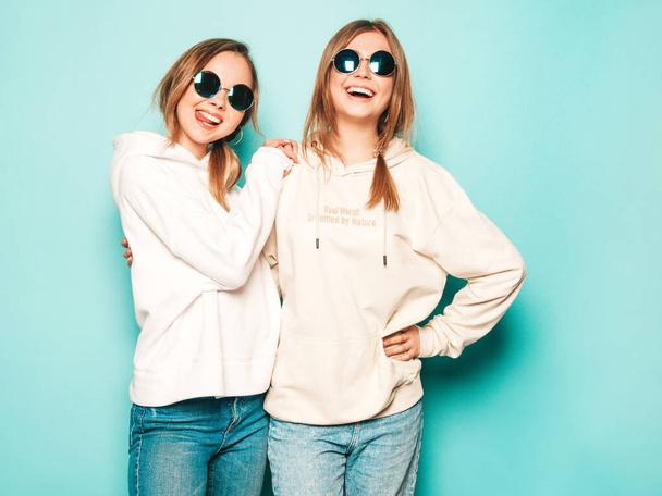 Two young beautiful blond smiling hipster girls in trendy summer hoodie clothes. Sexy carefree women posing near blue wall. Trendy and positive models having fun in sunglasses - Fotografie, Obrázek