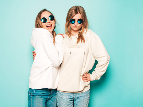 Two young beautiful blond smiling hipster girls in trendy summer hoodie clothes. Sexy carefree women posing near blue wall. Trendy and positive models having fun in sunglasses - Photo, Image