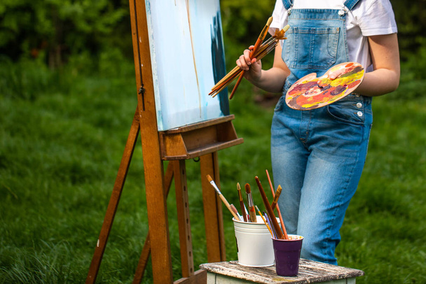 Μια νεαρή γυναίκα καλλιτέχνης κρατά μια βούρτσα και ζωγραφίζει μια εικόνα σε ένα καβαλέτο στις ακτίνες του ηλιοβασιλέματος. Ο ζωγράφος ζωγραφίζει ελαιογραφίες στο ηλιοβασίλεμα. - Φωτογραφία, εικόνα